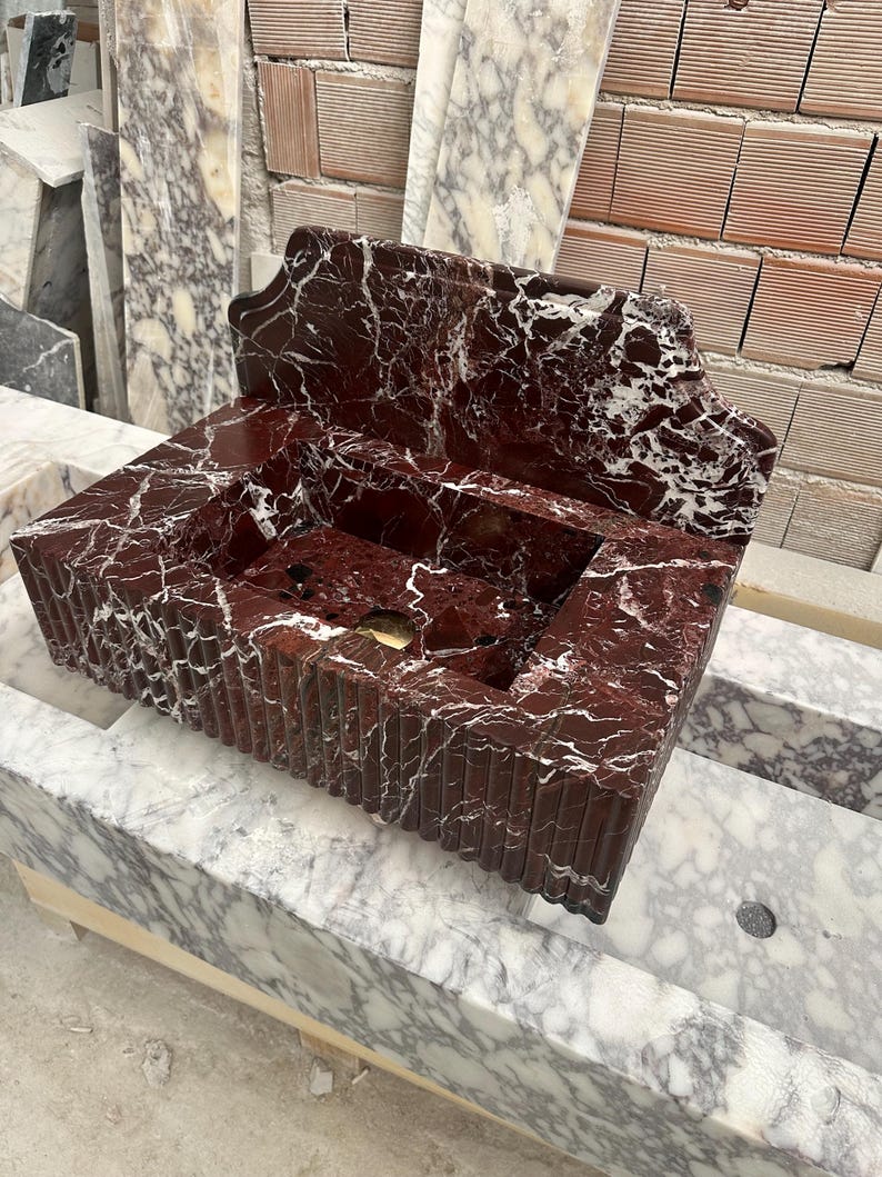 May include: A rectangular, red marble sink with white veining. The sink has a built-in backsplash and a grooved exterior design. The sink is made of stone and is a bathroom fixture.