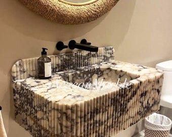 May include: A white marble bathroom sink with a wavy, fluted design. The sink is mounted on the wall and has a single basin. The sink is in a bathroom with a woven mirror and a woven wastebasket.