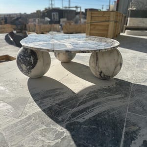 Puede incluir: Una mesa de centro de mármol con una tapa redonda y tres patas esféricas. La superficie de la mesa tiene un patrón veteado gris y blanco, y las patas son de un color similar. La mesa está diseñada para uso en interiores o exteriores.