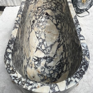 May include: A large, oval-shaped bathtub made of marble. The marble has a striking pattern of black, white, and beige swirls and veins. The tub has a smooth, polished finish and is designed for bathing. The overall aesthetic is luxurious and elegant.