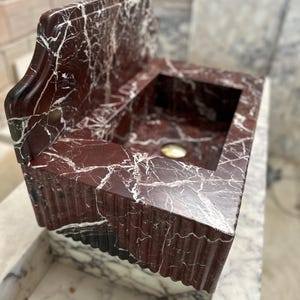 May include: A red marble sink with white veining. The sink has a rectangular basin and a backsplash. The front edge of the sink has a decorative, fluted design. The sink is made of a polished stone.