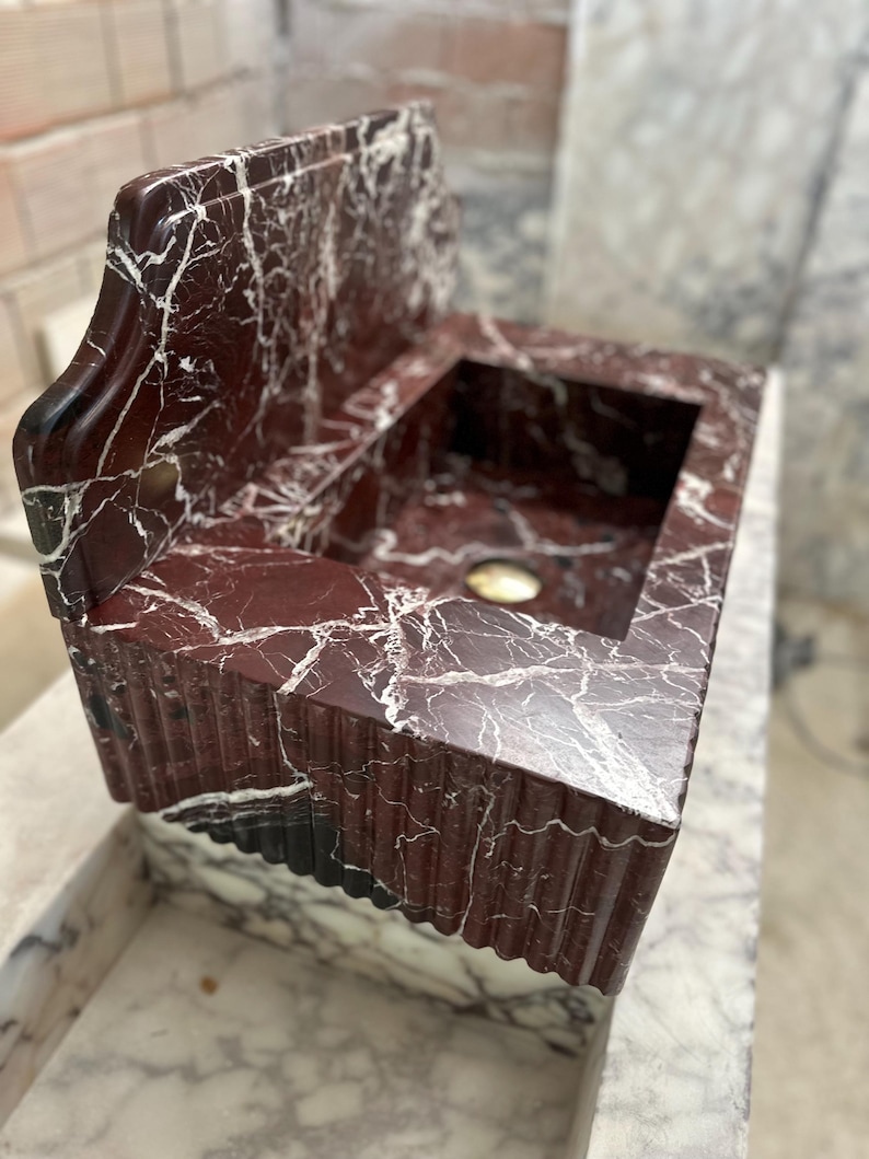 May include: A red marble sink with white veining. The sink has a rectangular basin and a backsplash. The front edge of the sink has a decorative, fluted design. The sink is made of a polished stone.