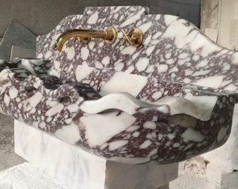 May include: A large, white marble sink with a wavy edge and brown and white marbling. The sink has a gold faucet and is mounted on a white marble pedestal.