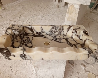 May include: A white marble sink with brown veining. The sink has a wavy edge and a round drain hole.