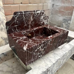 May include: A rectangular, red marble sink with white veining. The sink has a decorative back and a ribbed front. The sink is sitting on a marble surface. The sink is likely for a bathroom.