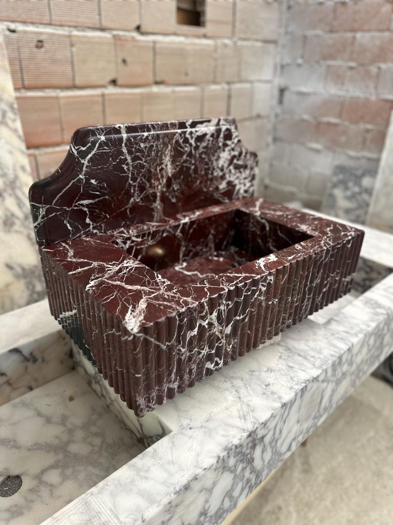May include: A rectangular, red marble sink with white veining. The sink has a decorative back and a ribbed front. The sink is sitting on a marble surface. The sink is likely for a bathroom.