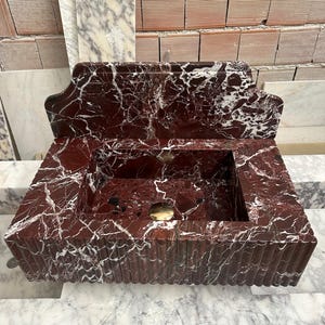 May include: A rectangular, red marble sink with white veining. The sink has a built-in backsplash and a fluted design on the front. A gold-colored drain is visible in the basin. The sink is made of a polished stone material.
