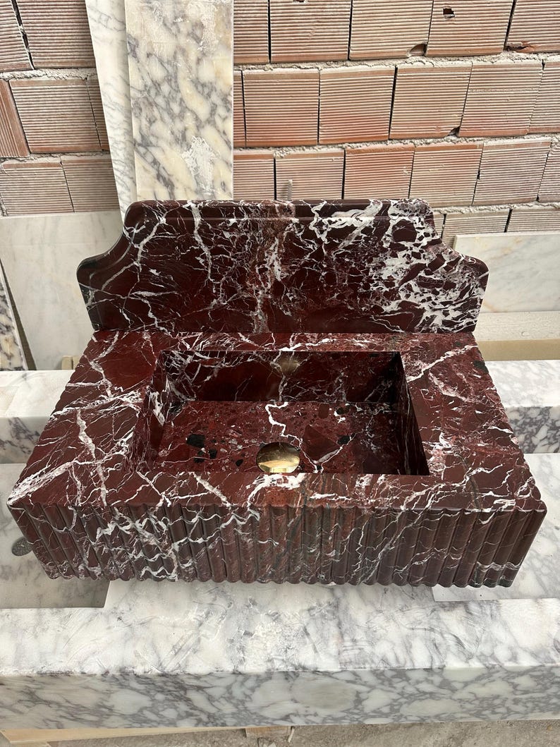 May include: A rectangular, red marble sink with white veining. The sink has a built-in backsplash and a fluted design on the front. A gold-colored drain is visible in the basin. The sink is made of a polished stone material.