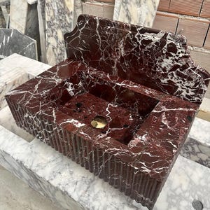 May include: A rectangular, red marble sink with white veining. The sink has a fluted front and a built-in backsplash. A gold-colored drain is visible in the center of the basin. The sink is made of natural stone.