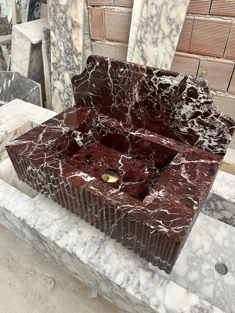 May include: A rectangular, red marble sink with white veining. The sink has a fluted front and a built-in backsplash. A gold-colored drain is visible in the center of the basin. The sink is made of natural stone.