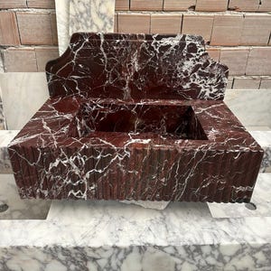 May include: A rectangular, red marble sink with white veining. The sink has a raised back and a rectangular basin. The front of the sink has vertical grooves. The sink is sitting on a marble surface.