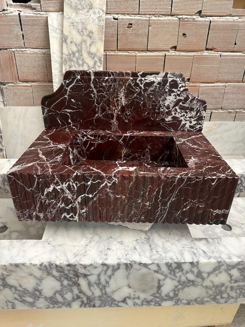 May include: A rectangular, red marble sink with white veining. The sink has a raised back and a rectangular basin. The front of the sink has vertical grooves. The sink is sitting on a marble surface.