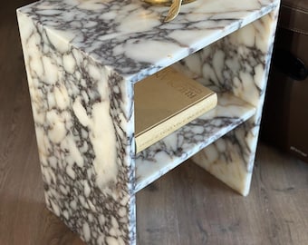 May include: A white marble end table with a gold whale figurine on top and a book on the lower shelf. The table has a unique open design with a shelf.