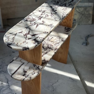 Puede incluir: Una mesa auxiliar moderna de dos niveles. La mesa presenta una encimera de mármol blanco con vetas marrones oscuras, sostenida por patas de madera natural. El diseño es minimalista y es adecuada para una sala de estar o dormitorio. Altura aproximada 76 cm.