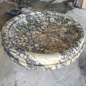 May include: A round marble sink with a speckled pattern of black and cream. The sink has a smooth, polished interior and a decorative rim. The sink has a small, centered drain hole.
