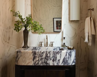 Puede incluir: Un mueble de baño con un lavabo de mármol con un patrón morado oscuro y blanco. El mueble tiene un mueble de madera oscura y un espejo con marco dorado. Dos apliques blancos flanquean el espejo y un jarrón con vegetación se encuentra en el lavabo.