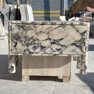 Puede incluir: Una mesita de noche de mármol blanco con un cajón y una base de madera. La mesita de noche tiene un patrón de mármol único con vetas negras y marrones.