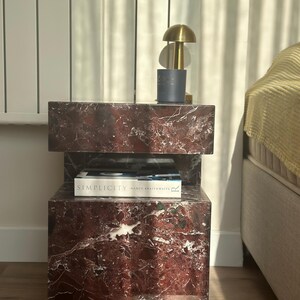 May include: A two-tiered red marble side table with a book titled "Simplicity" by Nancy Braithwaite on the top tier. A gold lamp with a white shade sits on the top tier.
