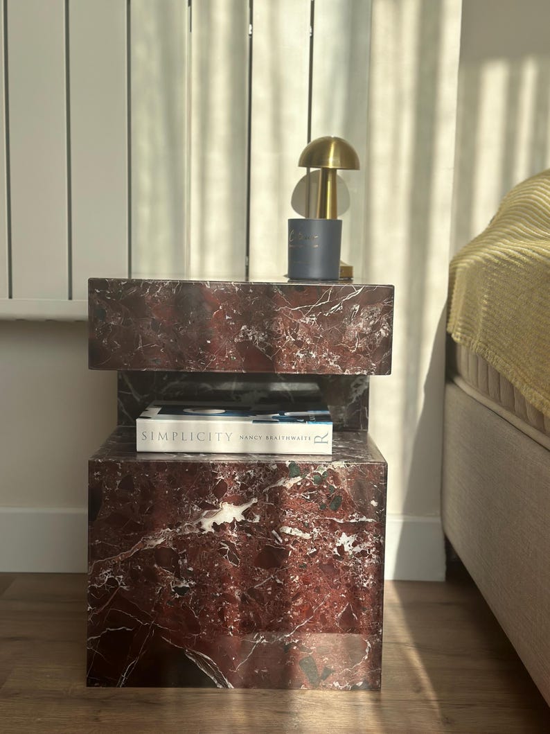 May include: A two-tiered red marble side table with a book titled "Simplicity" by Nancy Braithwaite on the top tier. A gold lamp with a white shade sits on the top tier.