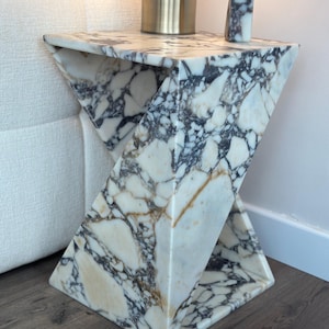Può includere: Un tavolino in marmo bianco con un design geometrico unico. Il tavolo presenta un sorprendente motivo di venature nere e marroni. Un oggetto cilindrico dorato e un piccolo blocco di marmo sono appoggiati sopra.