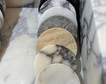 May include: A collection of round marble coasters in various colors and patterns, including white, gray, black, and beige. The coasters are arranged in a row, showcasing the natural veining and textures of the stone.