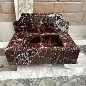 May include: A rectangular, red marble sink with white veining. The sink has a decorative back and a ridged front. The sink is sitting on a marble surface. The background is a brick wall.