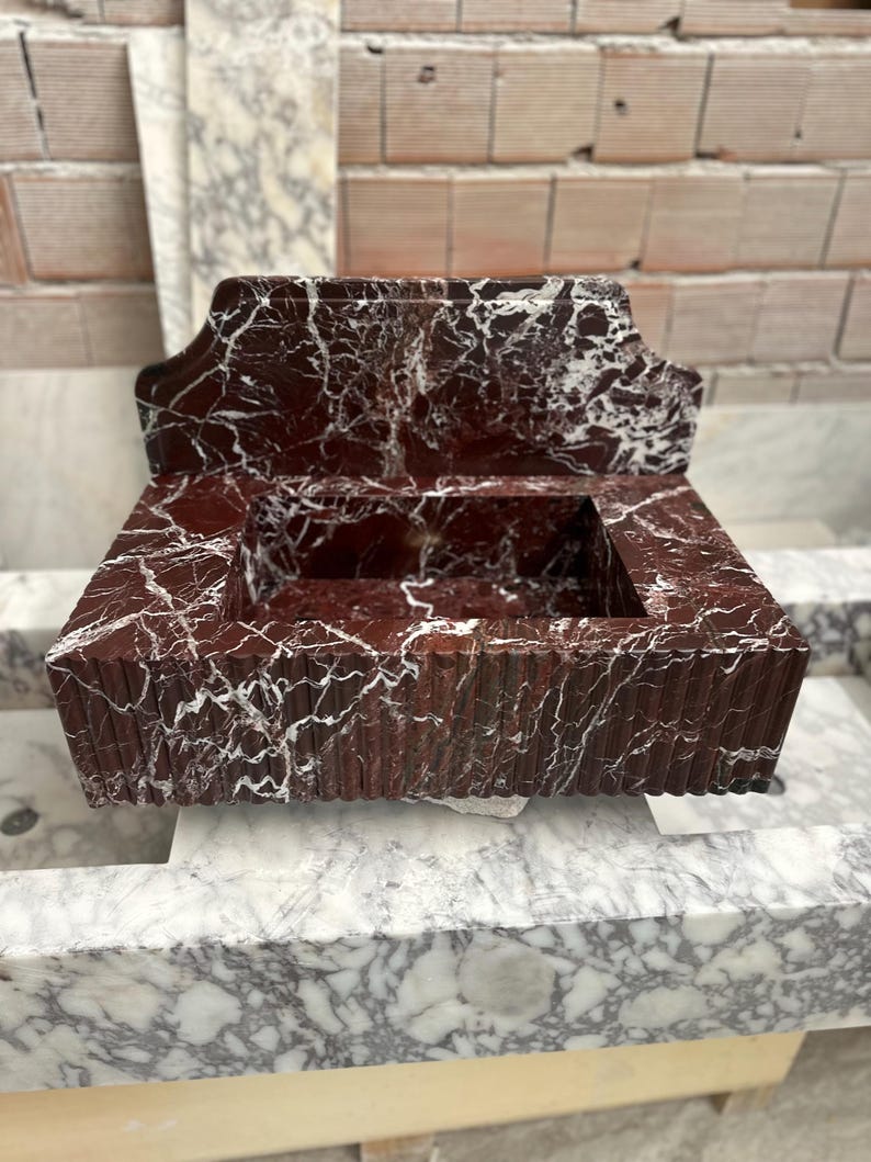 May include: A rectangular, red marble sink with white veining. The sink has a decorative back and a ridged front. The sink is sitting on a marble surface. The background is a brick wall.
