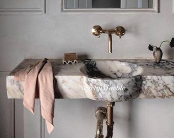 Può includere: Un lavabo da bagno in marmo con un lavabo integrato, con una miscela di venature bianche e grigie. Il lavabo ha un rubinetto e un sifone in ottone. Un asciugamano rosa è appeso di lato.