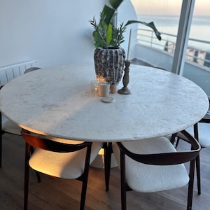 May include: A round, light-colored dining table with a marble-like surface, surrounded by four chairs with dark wooden frames and cream-colored seats. A decorative vase with greenery, candles, and a candlestick holder are on the table. The table is approximately 5 feet in diameter.