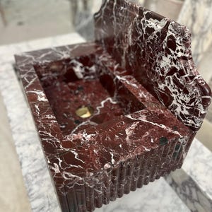 May include: A rectangular, red marble sink with white veining. The sink has a built-in backsplash and a fluted front. A gold-colored drain is visible in the basin. The sink is set on a marble surface.