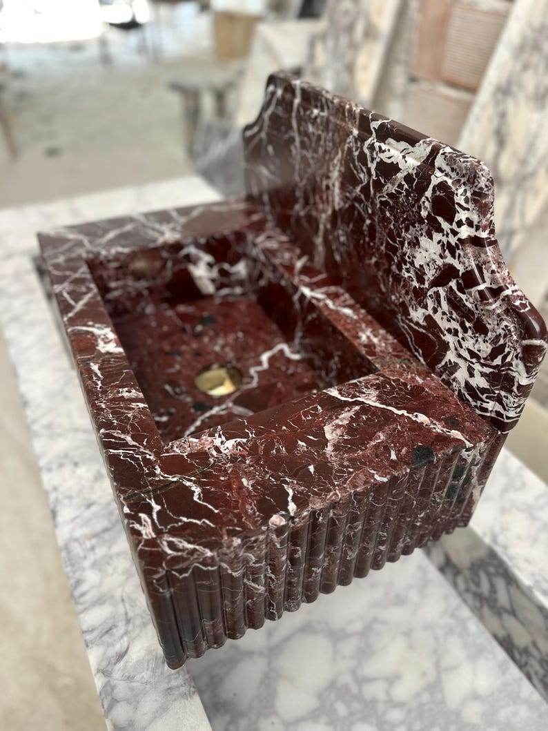 May include: A rectangular, red marble sink with white veining. The sink has a built-in backsplash and a fluted front. A gold-colored drain is visible in the basin. The sink is set on a marble surface.