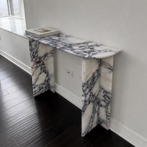 May include: A console table crafted from white marble with prominent dark grey and black veining. The table features a rectangular top with rounded edges and two vertical slab legs. A rectangular book rests on the left side of the table surface.