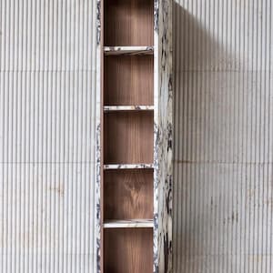 Op de afbeelding: Hoge, smalle houten kast met een deur met marmerpatroon. De kast heeft een donkerhouten interieur met meerdere planken. De deur is open en toont de planken en het interieur. De kast staat tegen een getextureerde, lichtgekleurde muur.