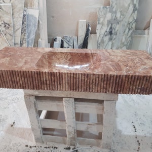 May include: A brown marble bathroom sink with a carved, fluted edge. The sink is mounted on a wooden stand.