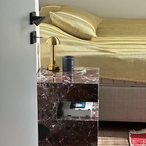 May include: A marble bedside table with a gold lamp and a book on the lower shelf. The table has a unique design with a cutout shelf.