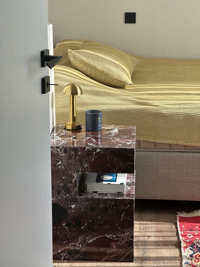 May include: A marble bedside table with a gold lamp and a book on the lower shelf. The table has a unique design with a cutout shelf.
