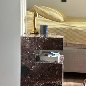 May include: A dark brown marble bedside table with a gold lamp and a blue candle on top. The table has a shelf underneath for storage.