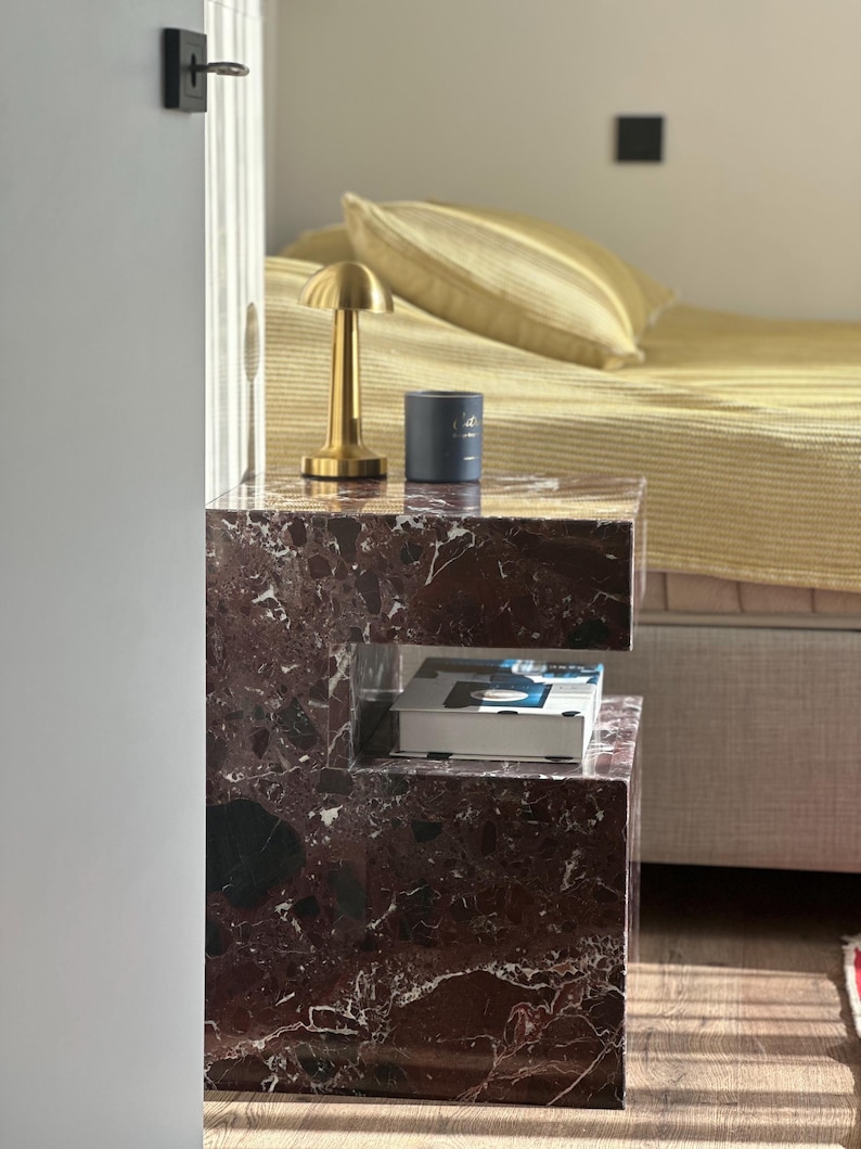 May include: A dark brown marble bedside table with a gold lamp and a blue candle on top. The table has a shelf underneath for storage.