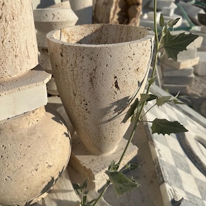 May include: A collection of beige stone planters in various shapes, including a large, textured, cone-shaped pot. Other planters are cylindrical and vase-like. Green leafy vines are draped across the planters, adding a natural element to the display.