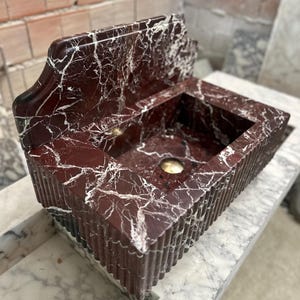 May include: A rectangular, red marble sink with white veining. The sink has a built-in backsplash and a decorative, fluted exterior. A gold-colored drain is visible in the basin. The sink is displayed on a marble surface.