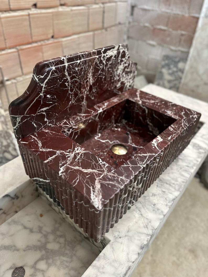 May include: A rectangular, red marble sink with white veining. The sink has a built-in backsplash and a decorative, fluted exterior. A gold-colored drain is visible in the basin. The sink is displayed on a marble surface.