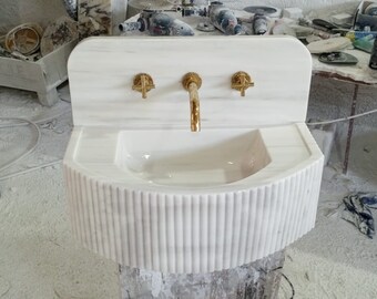 Op de afbeelding: Een witte marmeren wastafel met een gebogen ontwerp en gouden kranen. De wastafel is aan de muur gemonteerd en heeft een decoratieve, geribbelde basis.