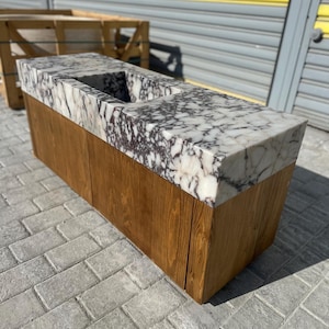 Puede incluir: Un mueble rectangular de madera con encimera de mármol. La encimera tiene un patrón blanco y gris y un recorte rectangular. El mueble es de madera marrón claro y se asienta sobre una superficie de ladrillo.