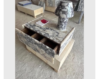 May include: A marble nightstand with a drawer open, revealing a wooden interior. A matching marble lamp sits on top, next to a small box. The nightstand is made of light beige and gray marble.