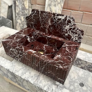 May include: A rectangular, red marble sink with white veining. The sink has a decorative back and a ridged front. A gold-colored drain is visible in the basin. The sink is set on a marble surface.
