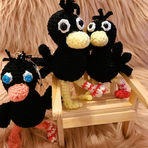 May include: Three black crocheted bird ornaments with white eyes and yellow beaks. The birds are sitting on a wooden bench with red and white striped legs.
