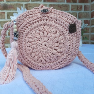 May include: A pink crocheted bag with a round, textured design and a long strap. The bag has a small, brown leather tag with the text "Handmade with Love" on it.