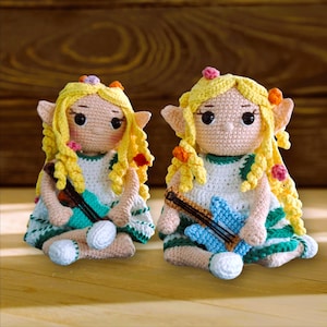 Peut inclure: Deux poupées crochetées à la main, avec des cheveux blonds bouclés, des oreilles pointues et tenant une guitare miniature. Les poupées portent des robes blanches avec des bordures vertes et des fleurs. Elles sont assises sur une surface en bois.