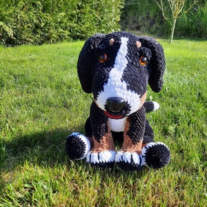 Könnte beinhalten: Ein gehäkeltes Stofftier in Form eines Hundes mit schwarzem, weißem und braunem Fell. Der Hund sitzt mit gekreuzten Pfoten und hat einen freundlichen Gesichtsausdruck.