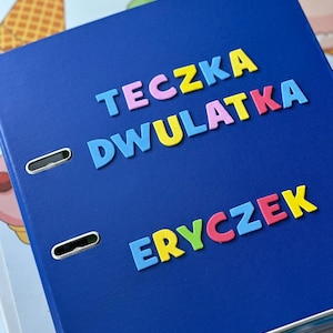 Może przedstawiać: Niebieski segregator z napisem "TECZKA DWULATKA ERYCZEK" w kolorowych literach.